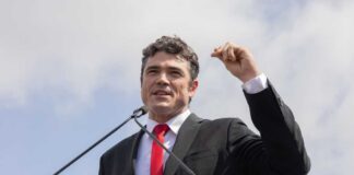 A man in a black suit and red tie speaking at a podium outdoors