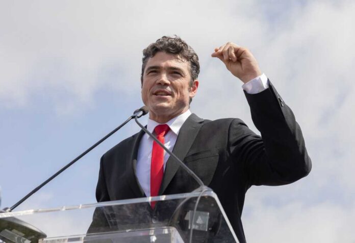 A man in a black suit and red tie speaking at a podium outdoors
