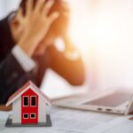 A businessman with his head in his hands, stressed over paperwork and a model house on the table