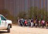 Group of children and adults walking near a border wall with a customs vehicle