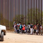 Group of children and adults walking near a border wall with a customs vehicle