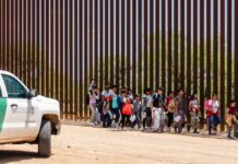 Group of children and adults walking near a border wall with a customs vehicle