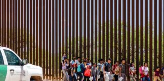 Group of children and adults walking near a border wall with a customs vehicle
