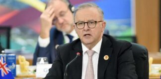 A man in a suit speaking at a conference table with a flag in the foreground