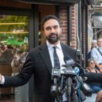 Man in a suit speaking at a press conference with microphones in front of him