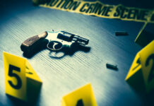 A handgun on a table with evidence markers and crime scene tape in the background