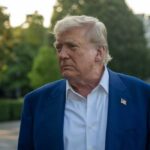 A political figure with a serious expression standing outdoors near the White House