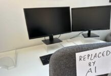 An office desk with two computer monitors and a chair labeled 'REPLACED BY AI'