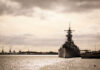 A naval ship docked in a harbor with industrial cranes in the background under a cloudy sky