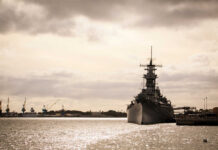 A naval ship docked in a harbor with industrial cranes in the background under a cloudy sky