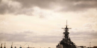 A naval ship docked in a harbor with industrial cranes in the background under a cloudy sky