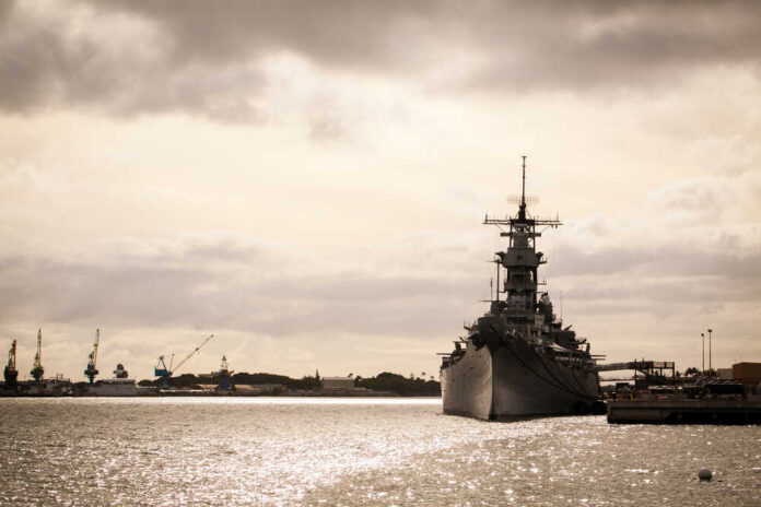 A naval ship docked in a harbor with industrial cranes in the background under a cloudy sky