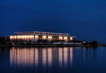 A modern building illuminated at night beside a calm body of water