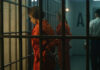 Inmates in orange jumpsuits stand in a jail corridor behind bars