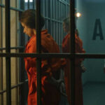 Inmates in orange jumpsuits stand in a jail corridor behind bars