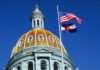 The dome of a state capitol building with flags flying against a blue sky