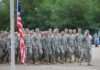 Group of soldiers in military uniforms marching in formation with an American flag in the foreground