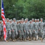 Group of soldiers in military uniforms marching in formation with an American flag in the foreground