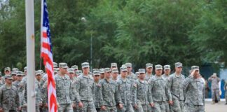 Group of soldiers in military uniforms marching in formation with an American flag in the foreground