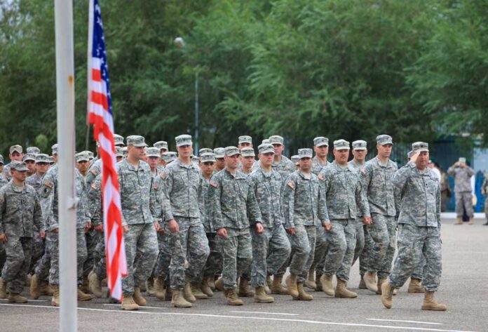 Group of soldiers in military uniforms marching in formation with an American flag in the foreground