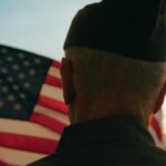 Emotional WWII Vet’s Plea: Remember Our Sacrifice A veteran in uniform standing in front of an American flag at sunset