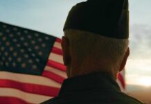 Emotional WWII Vet’s Plea: Remember Our Sacrifice A veteran in uniform standing in front of an American flag at sunset