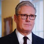 UK Politics in Turmoil: Reform’s Massive Gains A man in formal attire with glasses, looking serious in an indoor setting