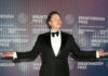 man in a tuxedo posing with arms outstretched at an awards event