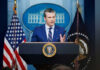 Man speaking at podium with flags in background