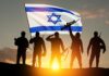 Silhouetted soldiers with rifles stand before an Israeli flag at sunset