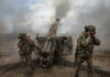 Soldiers fire a field howitzer as smoke and dust rise around them