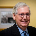 Questions Persist As Senator Recovers Smiling man in suit with round glasses indoors.