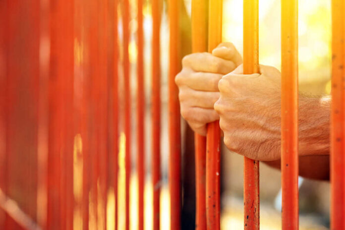Hands gripping orange prison bars with sunlight in the background