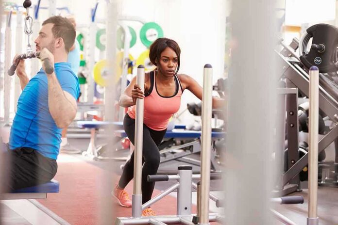 Woman performing sled push in a gym beside a man using a cable machine