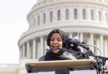 A Lawmaker’s Past Draws Renewed Federal Attention Woman in hijab speaking at podium with microphones