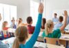 Students raise hands in a classroom with a teacher at the front