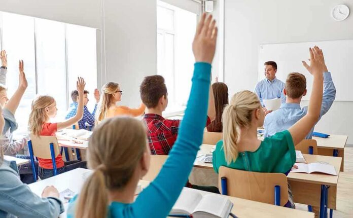 shutterstock_1470934406 Students raise hands in a classroom with a teacher at the front