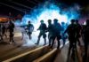 Ultra-Left Blamed As Cities Burn Riot police advance through tear gas at night on a city street