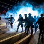 Ultra-Left Blamed As Cities Burn Riot police advance through tear gas at night on a city street