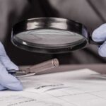 Gloved hands examine bullet casing with magnifying glass